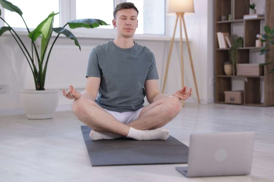 Un homme médite en position du lotus devant un ordinateur portable pour découvrir les bienfaits du yoga à domicile.
