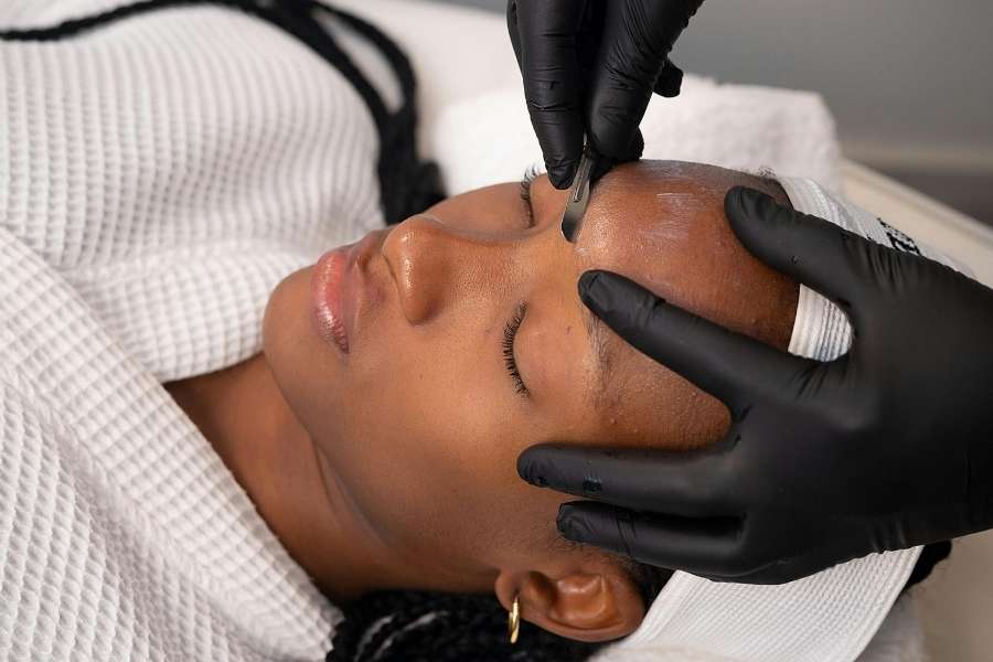 Esthéticienne en gants noirs réalisant un soin du visage réussi avec un instrument de dermaplaning sur le visage d'une cliente allongée.