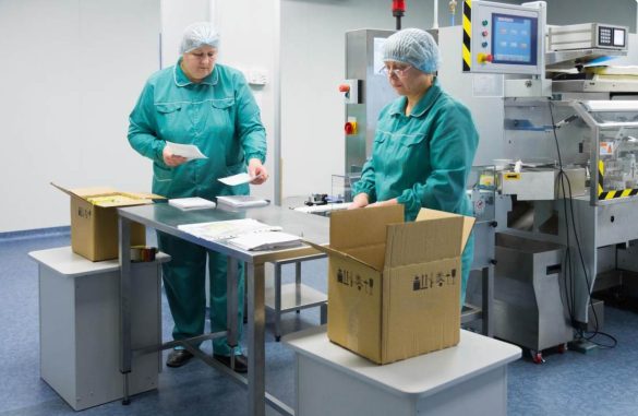 Deux techniciennes en blouse et charlotte gèrent l'emballage lors d'un déménagement de laboratoire près d'une machine de production.