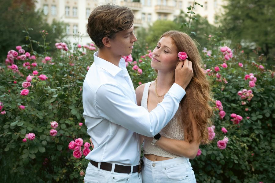 Un jeune homme en chemise blanche ajuste une rose dans les cheveux de sa compagne, illustrant une grande sensualité hors de la pénétration dans un jardin fleuri.